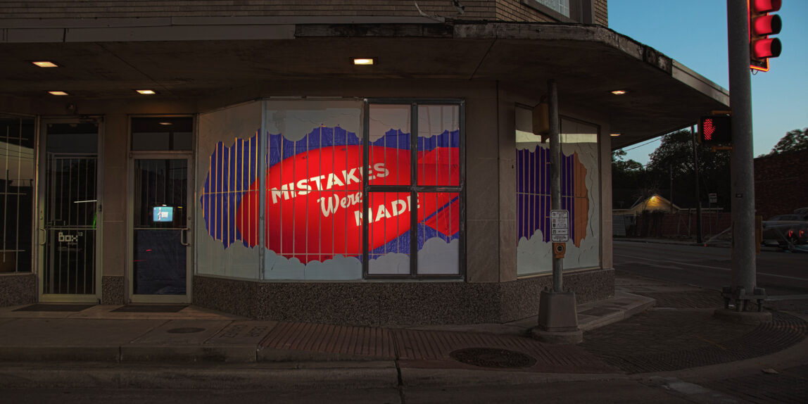 Large red inflatable balloon behind an old storefront window that has bars on the window. The words "Mistakes Were Made" are painted on the balloon and the balloon is shaped like a blimp. It is hung behind a backdrop of vivid blue tarps. The window is framed in layered white posterboard clouds.