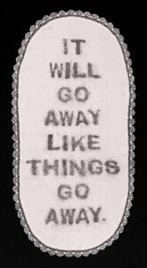 artwork displaying the remnants of charcoal soap letters on a white vintage tablecloth with lace border on a black background. The letters read "It will go away like things go away," a quote by Donald Trump.